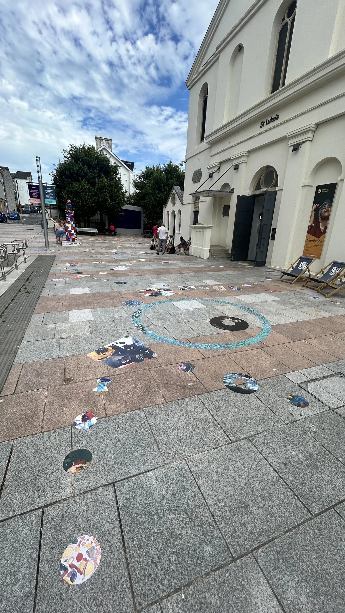 The finished courtyard in daylight, with cut out painting decals and a large teal ring scattered across the pink and grey paving in front of St Luke's