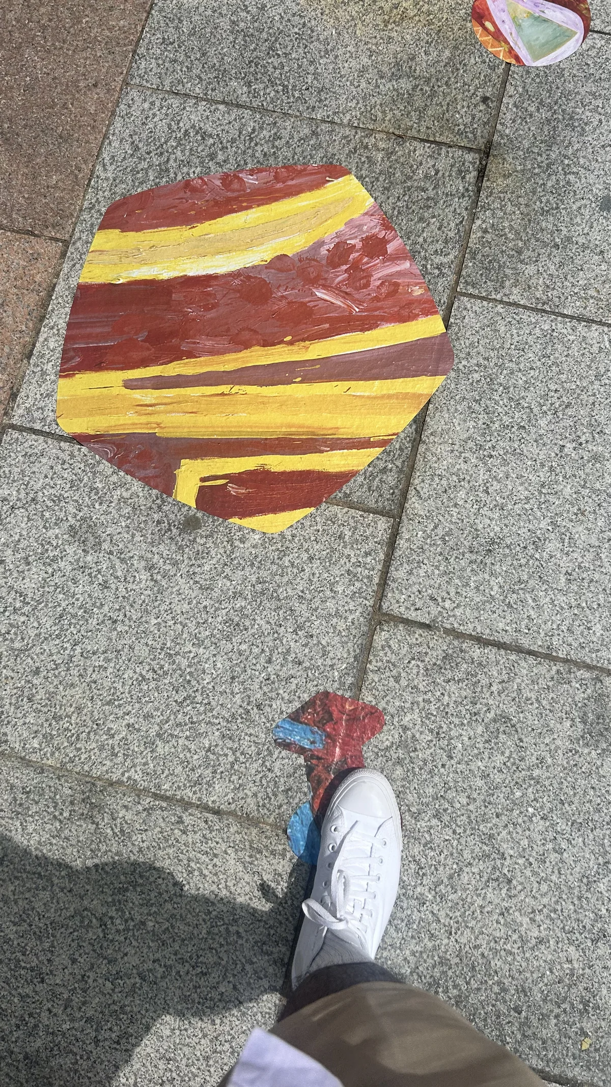 A red and yellow painted decal laid on grey granite paving, with a person's white trainer stepping towards a smaller red and blue shape