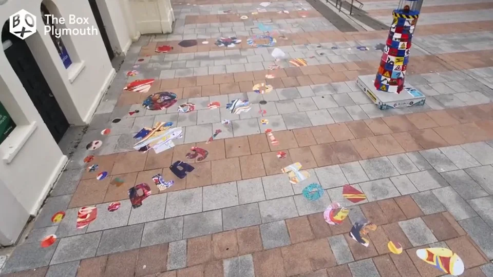 Two people on their knees laying painted floor decals across the paving outside The Box Plymouth, seen from above