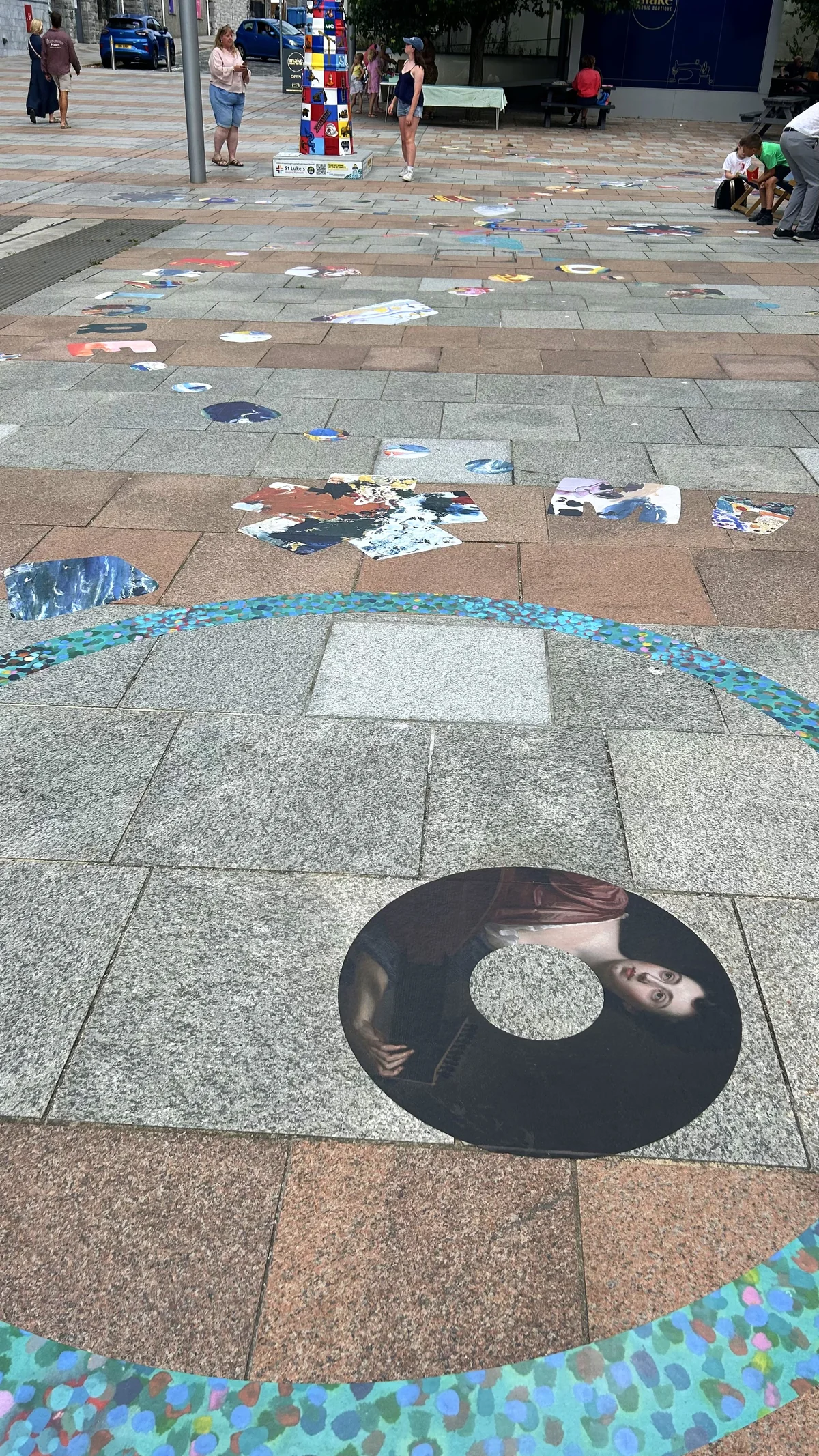 A black circle cut from a portrait of a woman playing a lute, on the paving, with a speckled teal ring curving around it and trails of decals beyond