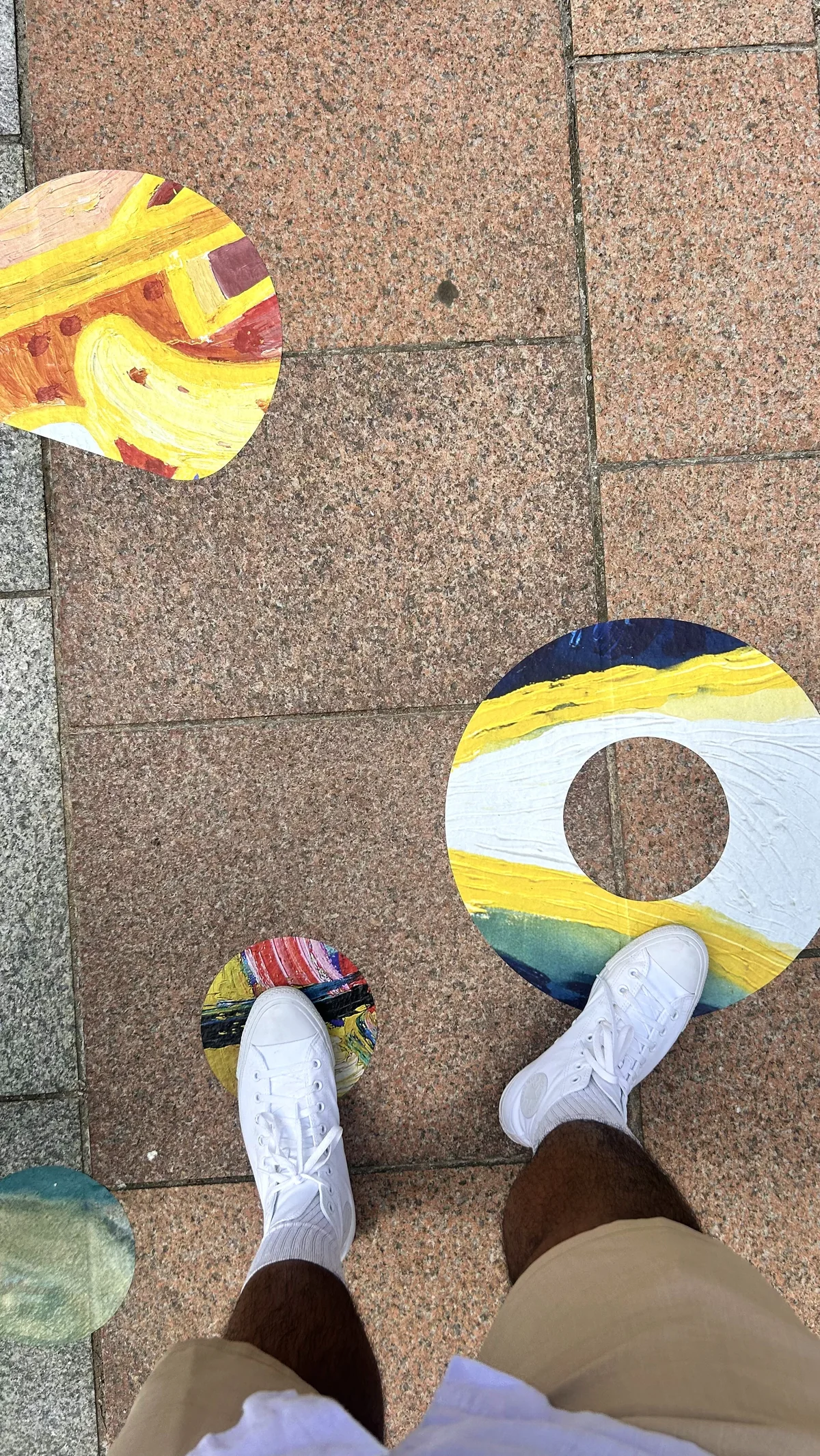 Three painted decals on pink paving, a yellow and white ring and two smaller shapes, with a person's trainers stepping between them