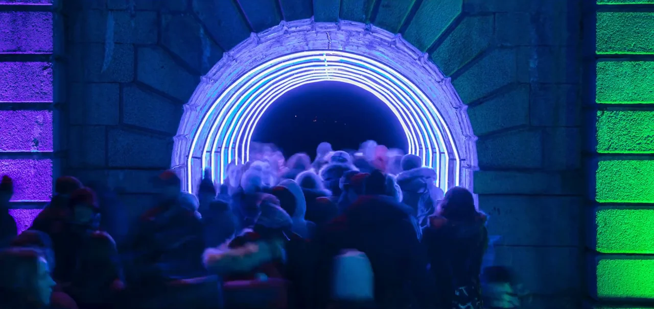 A timelapse of crowds in winter coats walking through the Freedom Tunnel, a stone arch lined with rings of blue and white light, beside walls lit purple and green