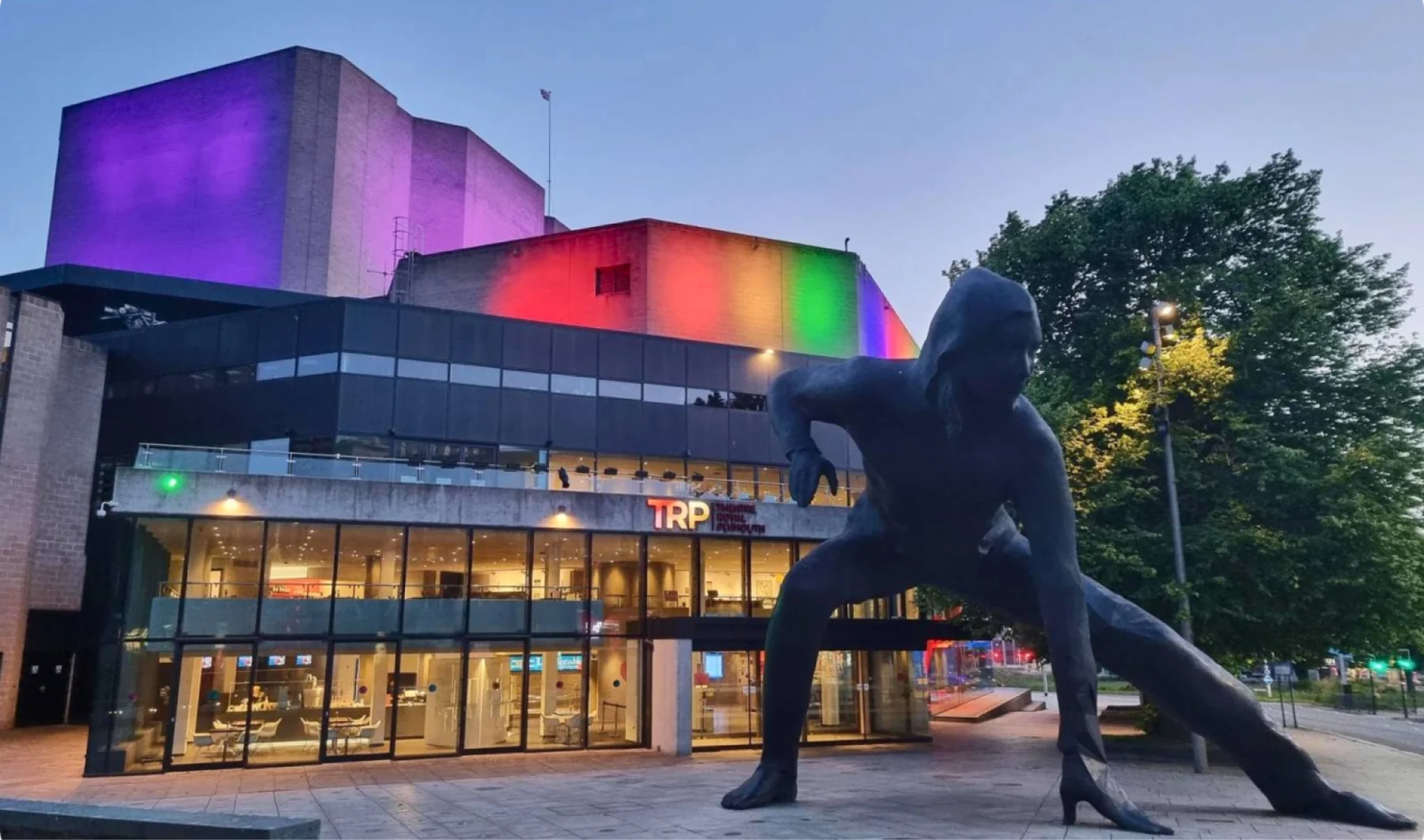 Theatre Royal Plymouth at dusk with its walls lit in purple, red, orange and green, and a large bronze figure of a crouching person in the foreground